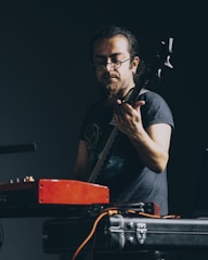 A musician focusing intently while playing an electric guitar, surrounded by equipment including a red amplifier and cables. The scene is dark, drawing attention to his focused expression and concentration on the music.