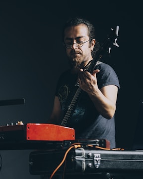 A musician focusing intently while playing an electric guitar, surrounded by equipment including a red amplifier and cables. The scene is dark, drawing attention to his focused expression and concentration on the music.