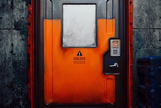 A sturdy, industrial-looking airlock door with an orange and gray color scheme positioned in a concrete wall. The door features a warning sign indicating the need to follow operational procedures. An access panel with a keypad and small display is mounted beside the door handle.