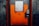 A sturdy, industrial-looking airlock door with an orange and gray color scheme positioned in a concrete wall. The door features a warning sign indicating the need to follow operational procedures. An access panel with a keypad and small display is mounted beside the door handle.