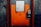 A sturdy, industrial-looking airlock door with an orange and gray color scheme positioned in a concrete wall. The door features a warning sign indicating the need to follow operational procedures. An access panel with a keypad and small display is mounted beside the door handle.