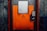 A sturdy, industrial-looking airlock door with an orange and gray color scheme positioned in a concrete wall. The door features a warning sign indicating the need to follow operational procedures. An access panel with a keypad and small display is mounted beside the door handle.