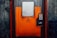 A sturdy, industrial-looking airlock door with an orange and gray color scheme positioned in a concrete wall. The door features a warning sign indicating the need to follow operational procedures. An access panel with a keypad and small display is mounted beside the door handle.