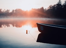 A serene lake at sunrise with a canoe anchored in the still water. Mist rises from the surface, and the sun casts a warm glow across the scene. Trees line the horizon, reflecting on the glassy water.