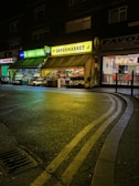 A night scene featuring a small supermarket with bright neon signs. The storefront is illuminated with green and yellow lights, showcasing an array of fruits and vegetables displayed outside. The street is wet, reflecting the vibrant lights, and a curved road with double yellow lines is visible in the foreground.