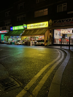A night scene featuring a small supermarket with bright neon signs. The storefront is illuminated with green and yellow lights, showcasing an array of fruits and vegetables displayed outside. The street is wet, reflecting the vibrant lights, and a curved road with double yellow lines is visible in the foreground.