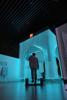 A person stands silhouetted within a large installation of vertical, glowing LED light strips. The lights are predominantly blue, giving the space a futuristic and immersive atmosphere. The ceiling is dark with visible beams, and there is a red exit sign above the installation. Information boards and signage are visible on stands in the background.
