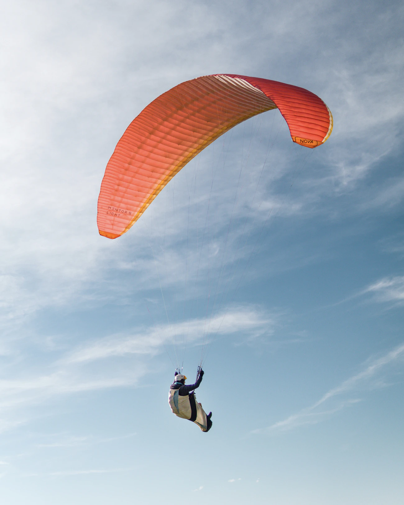 A pilot under an orange wing in a soft blue sky, harness clearly visible