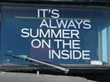 The image features a vehicle windshield with a printed phrase in bold white letters, 'IT'S ALWAYS SUMMER ON THE INSIDE.' A single windshield wiper is visible in the lower portion of the image, and reflections can be seen on the glass surface.