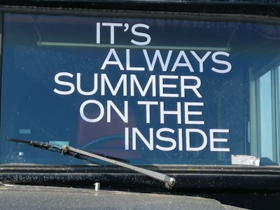 The image features a vehicle windshield with a printed phrase in bold white letters, 'IT'S ALWAYS SUMMER ON THE INSIDE.' A single windshield wiper is visible in the lower portion of the image, and reflections can be seen on the glass surface.