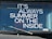 The image features a vehicle windshield with a printed phrase in bold white letters, 'IT'S ALWAYS SUMMER ON THE INSIDE.' A single windshield wiper is visible in the lower portion of the image, and reflections can be seen on the glass surface.