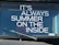 The image features a vehicle windshield with a printed phrase in bold white letters, 'IT'S ALWAYS SUMMER ON THE INSIDE.' A single windshield wiper is visible in the lower portion of the image, and reflections can be seen on the glass surface.