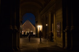 A nighttime view through an archway, revealing the illuminated glass pyramid of the Louvre Museum in the background. The scene includes people walking and standing under the arches, with warm ambient lighting from street lamps casting shadows on the stone architecture.
