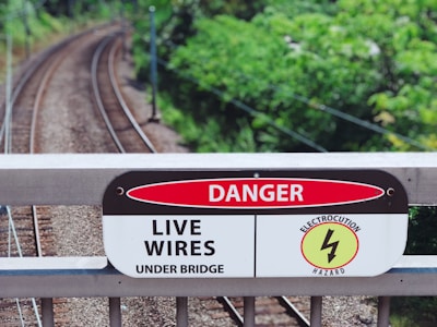 A railway track runs through a lush green area with a warning sign mounted on a bridge railing. The sign reads 'Danger: Live Wires Under Bridge' and features a symbol indicating an electrocution hazard.