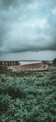 A lush green landscape is dominated by a large dam structure, with a vast reservoir of water extending into the distance. Above, a thick blanket of dark, moody clouds suggests an overcast or stormy atmosphere. The foreground is rich with dense vegetation, adding to the contrast between the natural environment and the man-made dam.