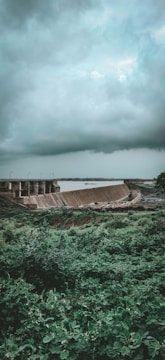 A lush green landscape is dominated by a large dam structure, with a vast reservoir of water extending into the distance. Above, a thick blanket of dark, moody clouds suggests an overcast or stormy atmosphere. The foreground is rich with dense vegetation, adding to the contrast between the natural environment and the man-made dam.