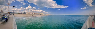 A panoramic view of a seaside amusement park and boardwalk. The scene includes a sandy beach with people sunbathing and playing, a boardwalk extending into the water, and a bright blue sky with scattered clouds. On the left, there is a distinctive tower and various park attractions.