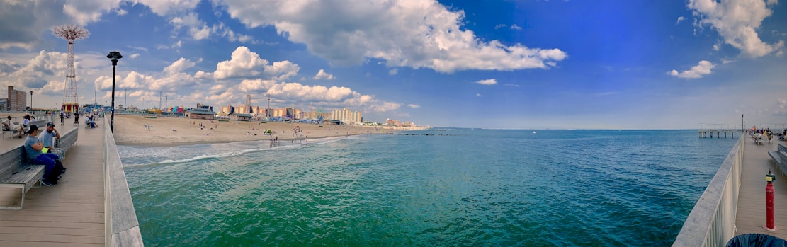 A panoramic view of a seaside amusement park and boardwalk. The scene includes a sandy beach with people sunbathing and playing, a boardwalk extending into the water, and a bright blue sky with scattered clouds. On the left, there is a distinctive tower and various park attractions.