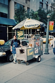 A street vendor stands by a hot dog cart on a city sidewalk. The cart is equipped with a large umbrella marked with 'Sabrett' and a sign advertising hot dogs and pretzels for a dollar. The surrounding area features a parked car, trees, and urban buildings, with a person walking nearby.