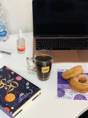 A workspace setup featuring an open laptop on a desk, with a glass mug filled with a dark beverage labeled Lipton in front of it. To the right, two doughnuts sit on a decorative purple napkin. On the left, there’s a colorful daily agenda book with a space-themed cover. A small bottle labeled 'ethyl alcohol' and a water bottle are also on the desk.