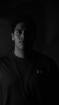A black and white portrait of a person with half of their face in shadow. The lighting creates a dramatic contrast, highlighting the features on one side and casting the other in darkness. The person is wearing a dark shirt with a logo visible.