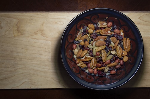 A wooden surface supports a dark ceramic bowl filled with various mixed nuts and dried fruits, including almonds, pecans, peanuts, raisins, and cranberries.