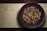 A wooden surface supports a dark ceramic bowl filled with various mixed nuts and dried fruits, including almonds, pecans, peanuts, raisins, and cranberries.