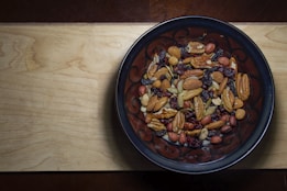 A wooden surface supports a dark ceramic bowl filled with various mixed nuts and dried fruits, including almonds, pecans, peanuts, raisins, and cranberries.