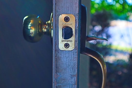 A close-up view of a door with a brass doorknob and a latch plate. The latch plate is engraved with the brand name Kwikset. The background is softly blurred with hints of greenery and light, suggesting an outdoor setting.
