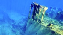 A submerged shipwreck rests on the ocean floor, surrounded by deep blue water. The ship's hull is partially intact, with visible metal structures and scattered debris. The scene evokes a sense of stillness and mystery beneath the sea.