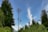 A tall communication tower is surrounded by lush green trees under a clear blue sky. Wispy clouds are visible in the background, adding texture to the sky. The scene conveys a mix of nature and technology.