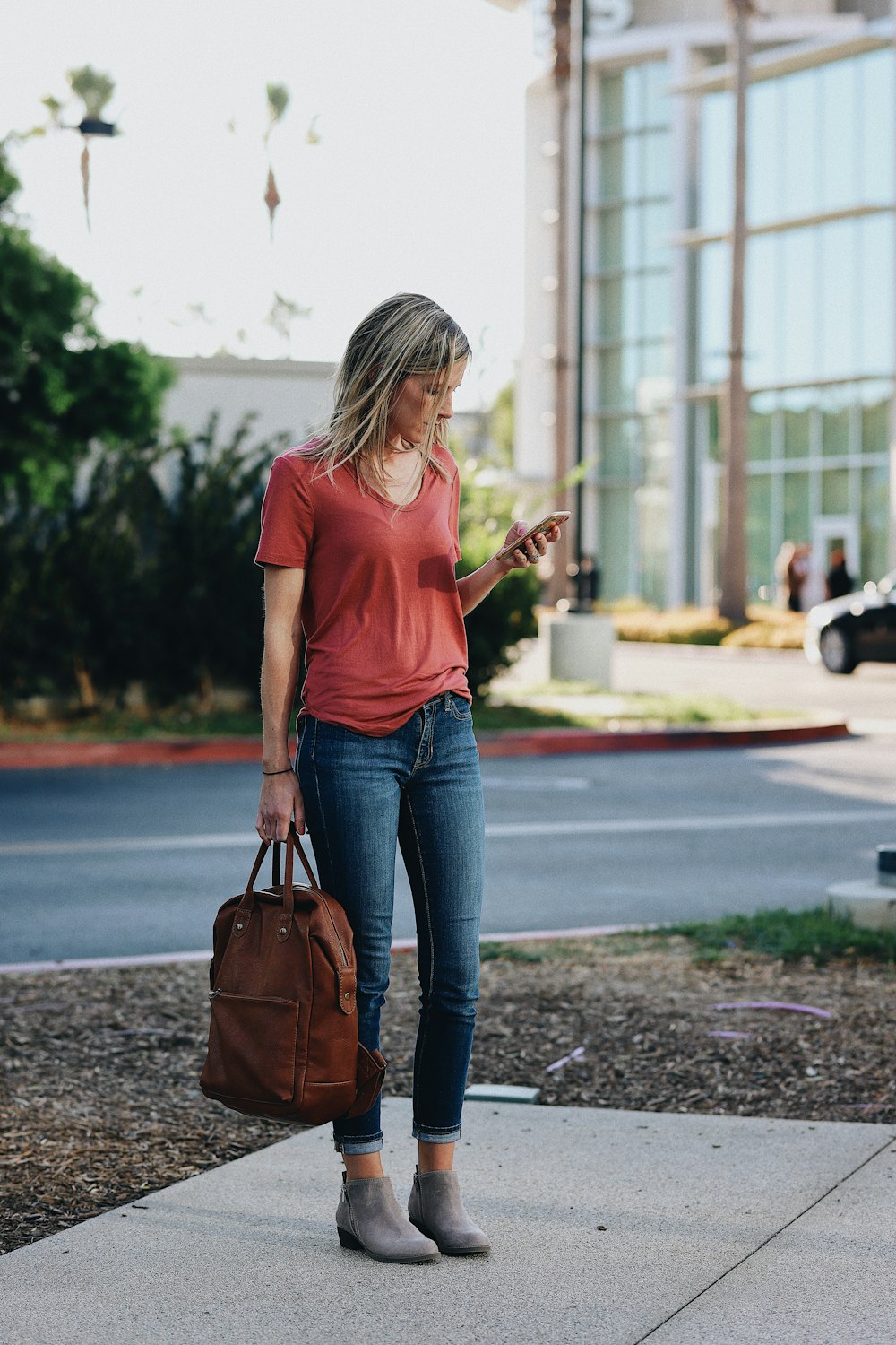 woman in red t-shirt and blue denim jeans holding brown leather sling bag