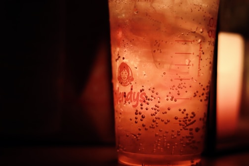 A close-up of a glass filled with a fizzy beverage featuring ice cubes and bubbles. The lighting creates a warm, amber glow, and the glass bears a logo. The ambiance is cozy and dimly lit.