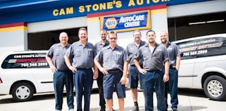 A group of seven men wearing matching blue work uniforms stands in front of a garage with signs denoting 'CAM STONE'S AUTOMOTIVE' and 'NAPA AutoCare Center.' Two white vans with signage promoting auto services are visible. The men appear to be staff members or mechanics.