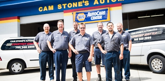 A group of seven men wearing matching blue work uniforms stands in front of a garage with signs denoting 'CAM STONE'S AUTOMOTIVE' and 'NAPA AutoCare Center.' Two white vans with signage promoting auto services are visible. The men appear to be staff members or mechanics.