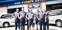 A group of seven men wearing matching blue work uniforms stands in front of a garage with signs denoting 'CAM STONE'S AUTOMOTIVE' and 'NAPA AutoCare Center.' Two white vans with signage promoting auto services are visible. The men appear to be staff members or mechanics.