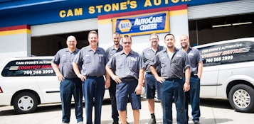 A group of seven men wearing matching blue work uniforms stands in front of a garage with signs denoting 'CAM STONE'S AUTOMOTIVE' and 'NAPA AutoCare Center.' Two white vans with signage promoting auto services are visible. The men appear to be staff members or mechanics.