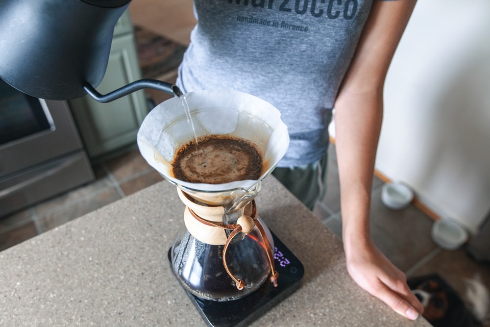 person in gray t-shirt pouring brown liquid on clear glass cup