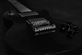 A close-up view of an electric guitar focusing on the bridge, pickup, and control knobs. The guitar has a sleek design with a glossy finish, and the image is in black and white, emphasizing the metal components and string details.