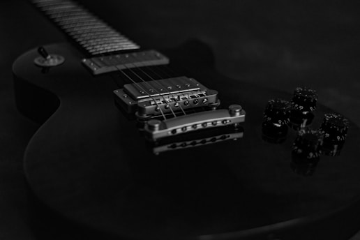 A close-up view of an electric guitar focusing on the bridge, pickup, and control knobs. The guitar has a sleek design with a glossy finish, and the image is in black and white, emphasizing the metal components and string details.