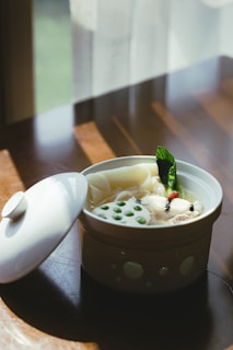 A ceramic bowl filled with a soup featuring sliced lotus root, green peas, leafy greens, and floating ingredients. The bowl has a lid placed slightly to the side on a wooden table. Sunlight streams through a nearby window, casting soft shadows.