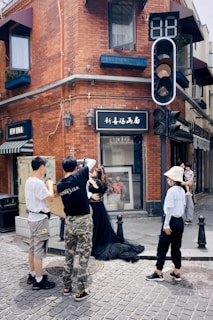 A bustling urban street corner with red brick buildings and a group of people engaged in a photoshoot. A person dressed in a dark elegant gown is being adjusted by another while two others observe. A traffic light and digital countdown are visible, and the scene includes storefronts with signage.