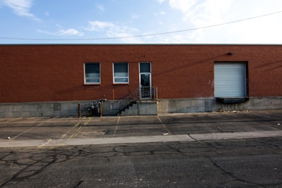 A large brick warehouse building with two small windows and a large garage door on the right. A concrete loading dock with stairs and a metal railing is visible, as well as a paved parking area with yellow lines and some cracks on the asphalt. Above, the sky is partly cloudy.