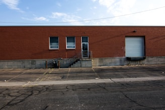 A large brick warehouse building with two small windows and a large garage door on the right. A concrete loading dock with stairs and a metal railing is visible, as well as a paved parking area with yellow lines and some cracks on the asphalt. Above, the sky is partly cloudy.