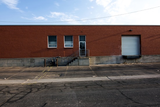 A large brick warehouse building with two small windows and a large garage door on the right. A concrete loading dock with stairs and a metal railing is visible, as well as a paved parking area with yellow lines and some cracks on the asphalt. Above, the sky is partly cloudy.