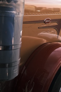 The image displays a close-up of a section of a large vehicle, likely a truck. A polished metallic exhaust pipe features prominently on the left, with a brown fender showing decorative details on the right. The vehicle's surface is smooth and reflective, with an emblem visible.