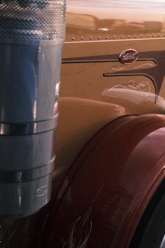 The image displays a close-up of a section of a large vehicle, likely a truck. A polished metallic exhaust pipe features prominently on the left, with a brown fender showing decorative details on the right. The vehicle's surface is smooth and reflective, with an emblem visible.