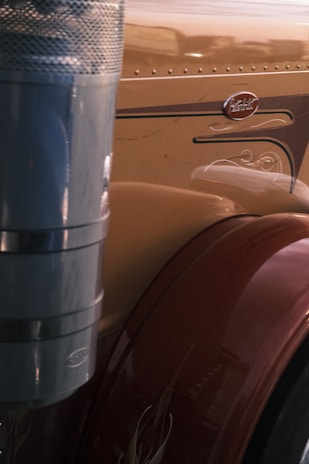 The image displays a close-up of a section of a large vehicle, likely a truck. A polished metallic exhaust pipe features prominently on the left, with a brown fender showing decorative details on the right. The vehicle's surface is smooth and reflective, with an emblem visible.