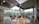 A modern, spacious lounge area with large ceiling-to-floor windows allowing plenty of natural light. The interior features a combination of gray sofas, bean bags, and small round tables on a wooden floor. The high ceiling has visible ductwork and lighting fixtures. The glass windows offer a view of greenery and a modern building exterior.