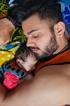 A man with a beard is lying down, peacefully sleeping with a newborn baby resting on his chest. They are both asleep, conveying a sense of calm and tenderness. The baby is wrapped in a colorful blanket, and the background is a mix of vibrant patterned fabrics.